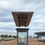 Parada de ônibus isolada com fita policial no Distrito Federal, cena de investigação sem pessoas, arquitetura de Brasília ao fundo.